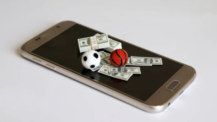 A smartphone with money, a soccer ball, and a basket ball on top of it. Gambling addiction is a serious condition, but therapy can help.