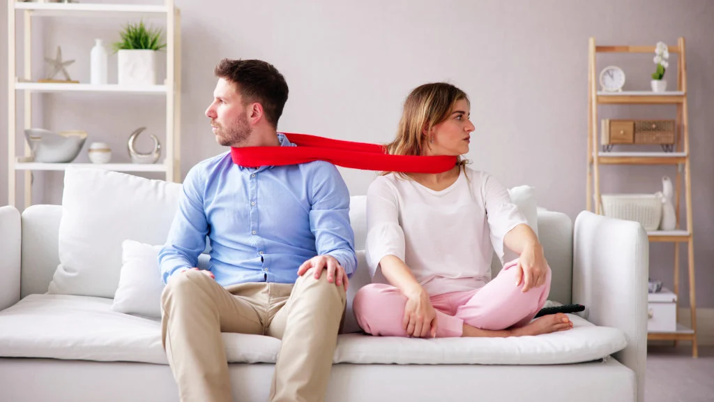 A couple sits on a couch, looking away from each other, with a scarf connecting them both